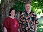 vor einem Baum, links eine Frau mit schulterlangen braunen Haaren und roter Bluse und ein Mann mit kurzen grauen Haaren und gemustertem Polo-Shirt